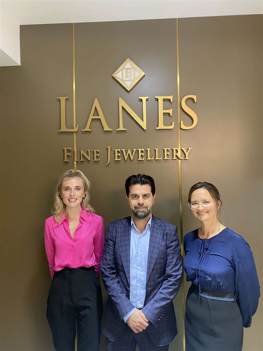 Ercan and Carli Onguc with sales associate Sophie Hatfield, standing beneath the gold Lanes Fine Jewellery wordmark inside the Red Lion Square showroom, Stamford